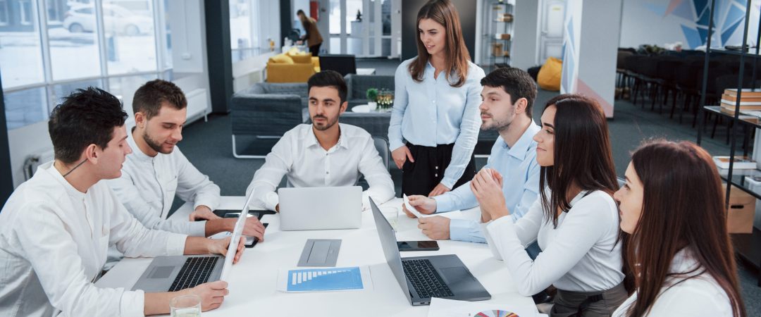 Guy on the left side talking and colleagues is listening to him. Group of young freelancers in the office have conversation and smiling.